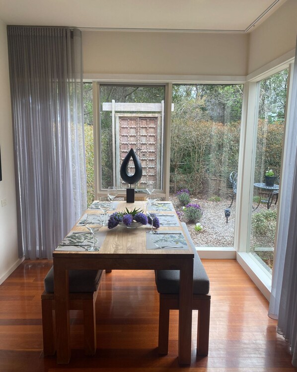 A dining area with a table set for four, a wooden bench, and a view of a garden through large windows.