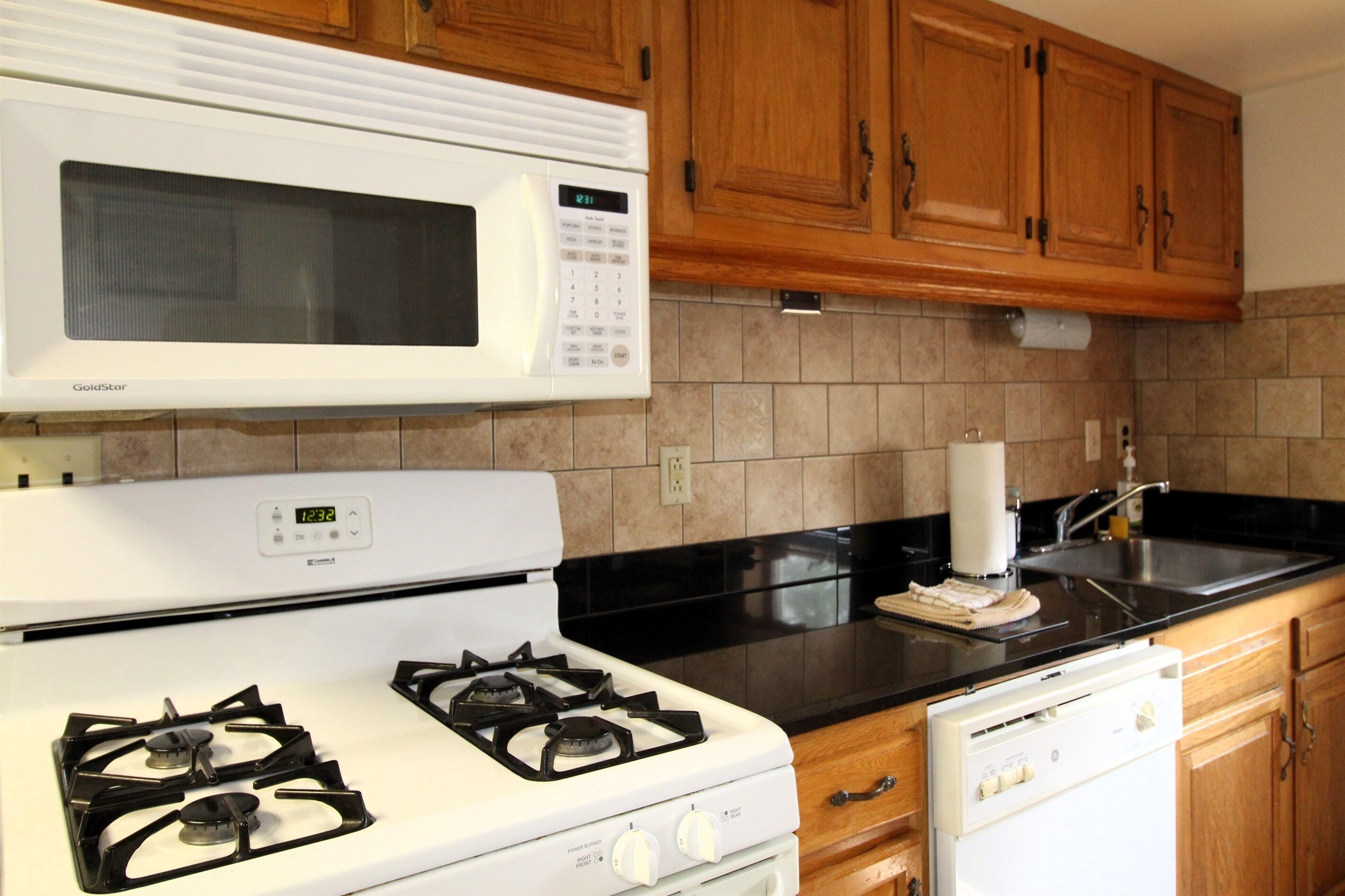 A kitchen with a white stove, a microwave, wooden cabinets, a black countertop, and a dishwasher.