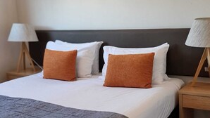Un letto rifatto con cuscini bianchi e marroni, una coperta grigia e un comodino in legno con una lampada.