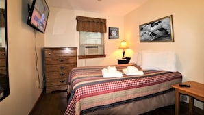 A hotel room with a bed, a dresser, a television, a window with curtains, a wall-mounted air conditioner, a framed picture, and a bedside lamp. - Cooperstown Baseball Rentals - Championship #1 (Oneonta)