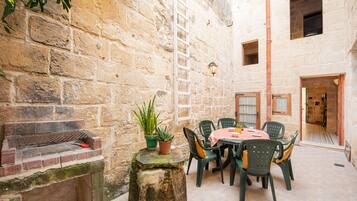 A stone-walled courtyard with a brick oven, a table with chairs, and potted plants.