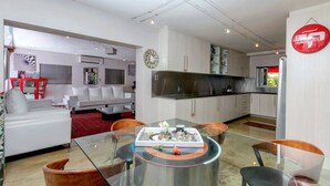 Uma sala de estar moderna com uma mesa de jantar de vidro, sofás brancos e uma área de cozinha com armários brancos e bancada preta.