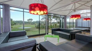 Espaço interior moderno com vista para uma área gramada. Apresenta assentos pretos, uma mesa verde e luminárias pendentes vermelhas.