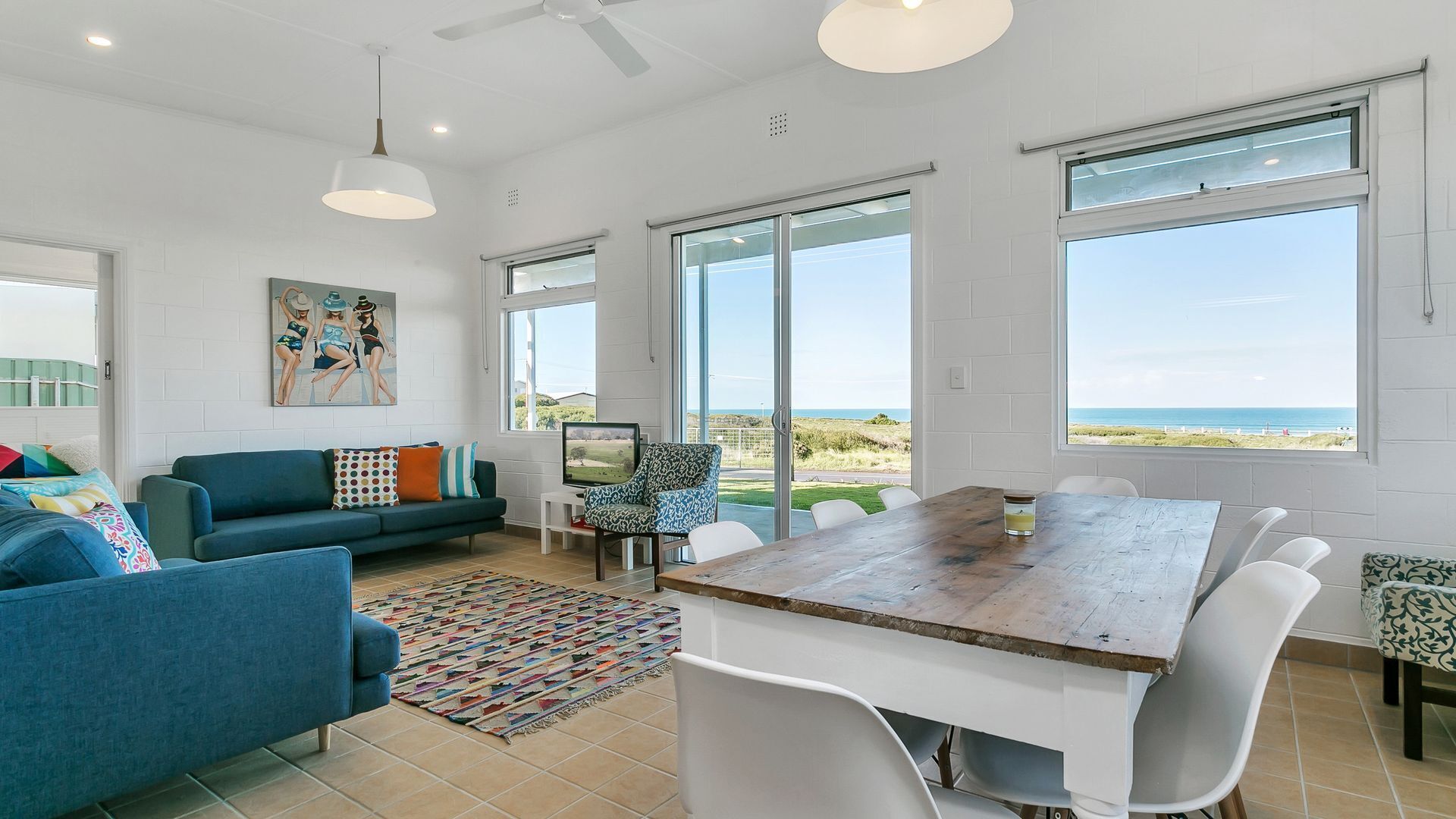A spacious living area with a large wooden table, white chairs, a blue sofa, and a colorful rug.