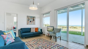 A modern living room with a blue sofa, colorful pillows, a patterned rug, and a large window offering a view of the beach.