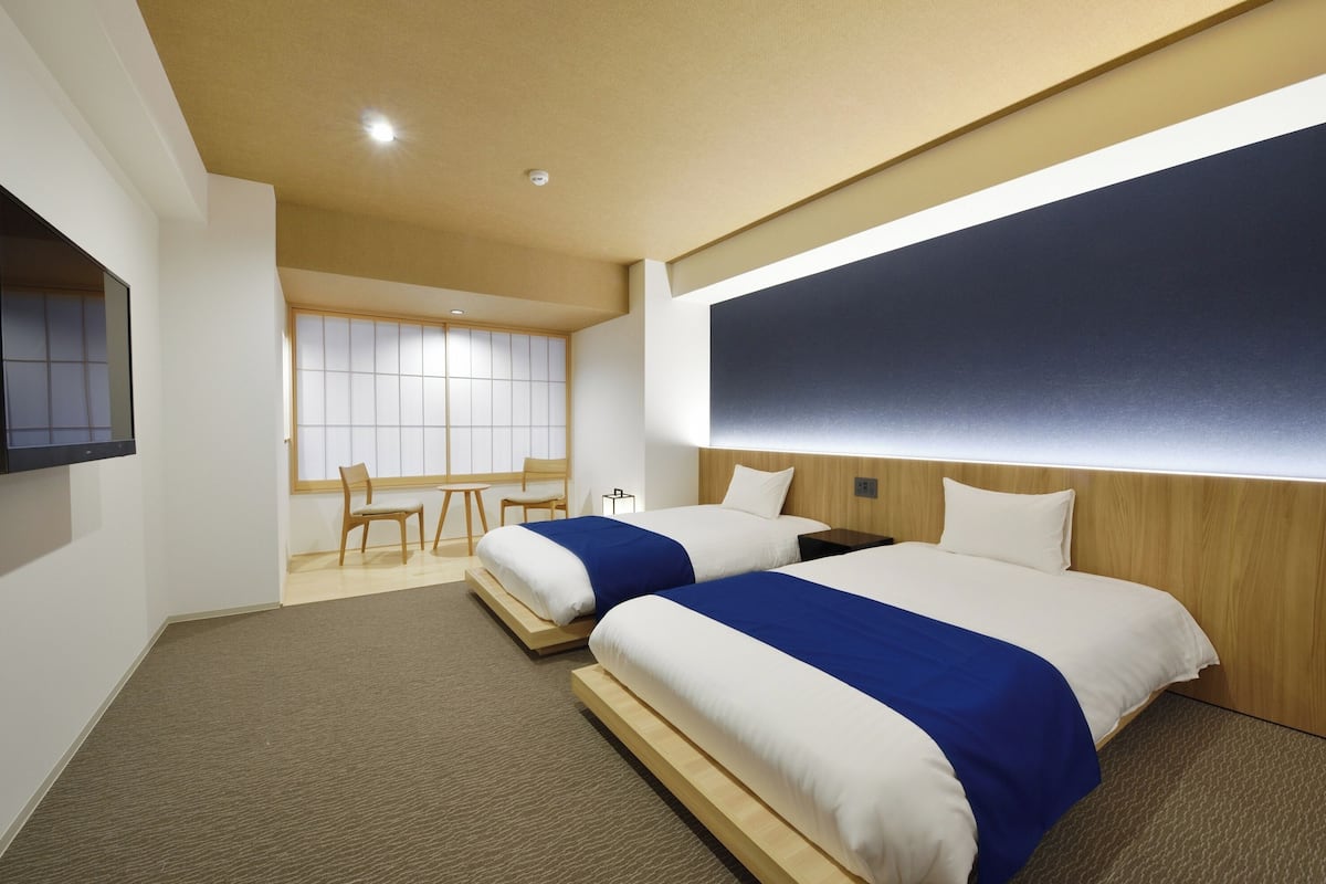 A hotel room with two beds, a TV, a wooden headboard, and a large window with blinds.