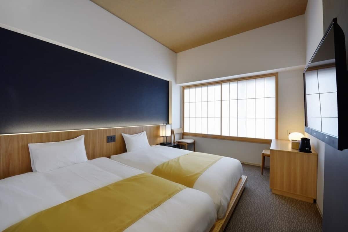 A hotel room with two beds, a large window, a desk, and a flat-screen TV.