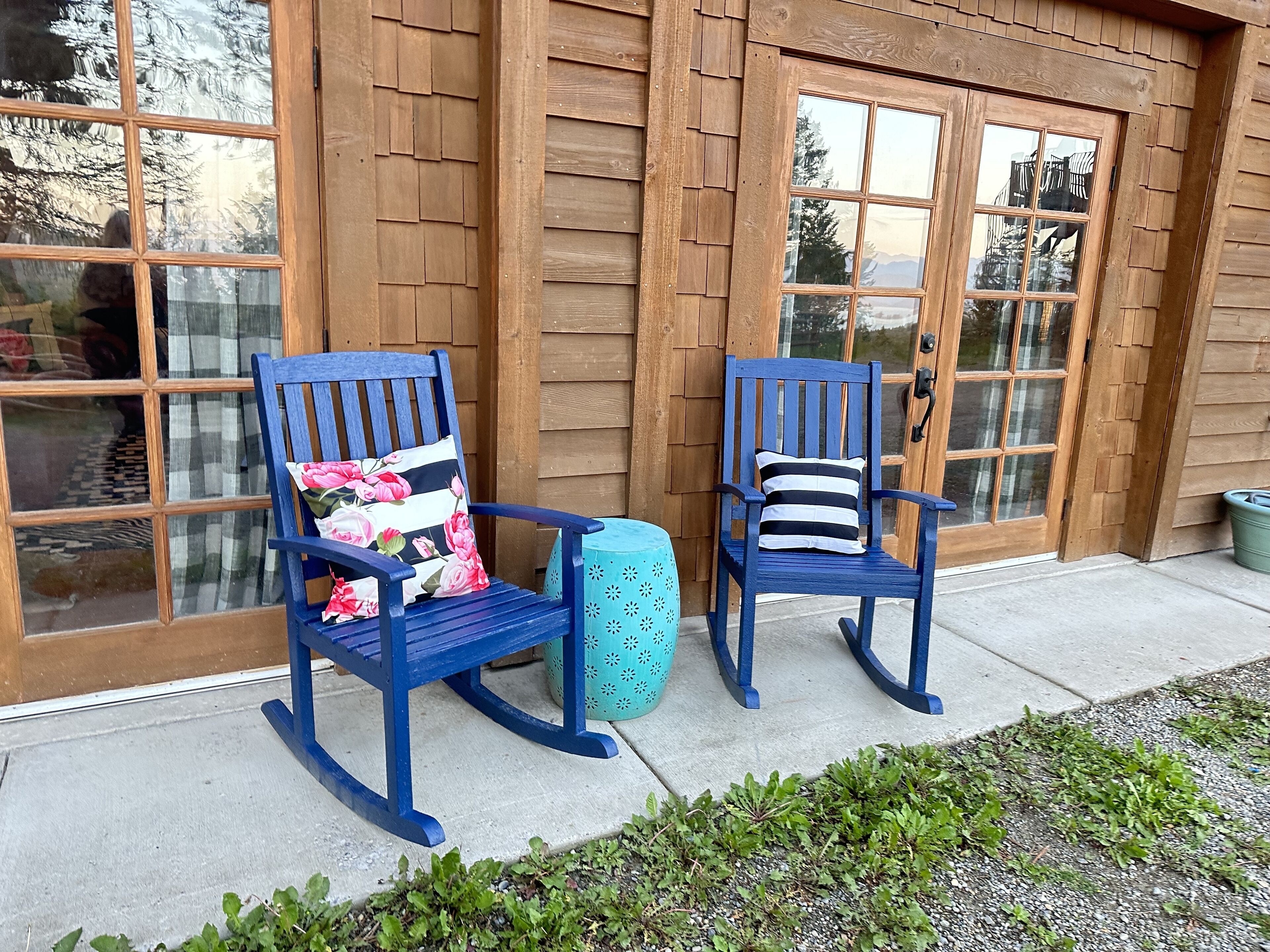 Two rocking chairs with floral and striped cushions, a turquoise stool, and a wooden building with large windows and doors.