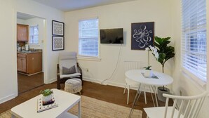 Ett kompakt bostadsutrymme med ett litet kök, en matplats med ett vitt bord och stolar, en sittgrupp med en bekväm fåtölj och ett stort fönster med persienner.