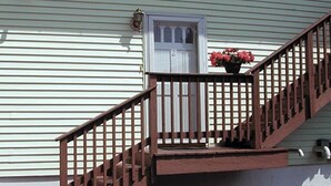 Une maison avec un escalier en bois menant à une porte blanche, un pot de fleurs contenant des fleurs roses et un petit jardin.
