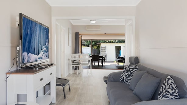 Ein modernes Wohnzimmer mit Fernseher, grauem Sofa und Essbereich mit weißen Stühlen.