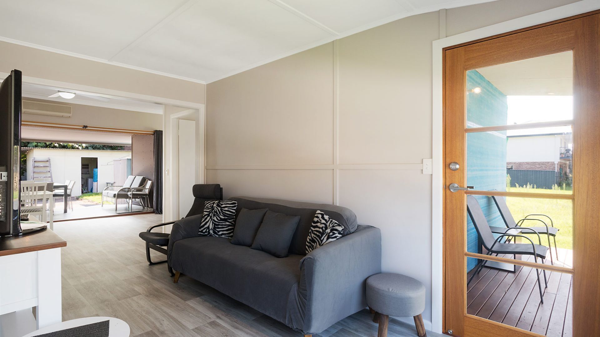 Un salon moderne avec un canapé gris, un petit pouf et un téléviseur posé sur un meuble. On y trouve une porte en bois avec un panneau de verre, offrant une vue sur l’extérieur.