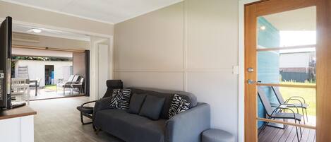 Un salon moderne avec un canapé gris, un petit pouf et un téléviseur posé sur un meuble. On y trouve une porte en bois avec un panneau de verre, offrant une vue sur l’extérieur.