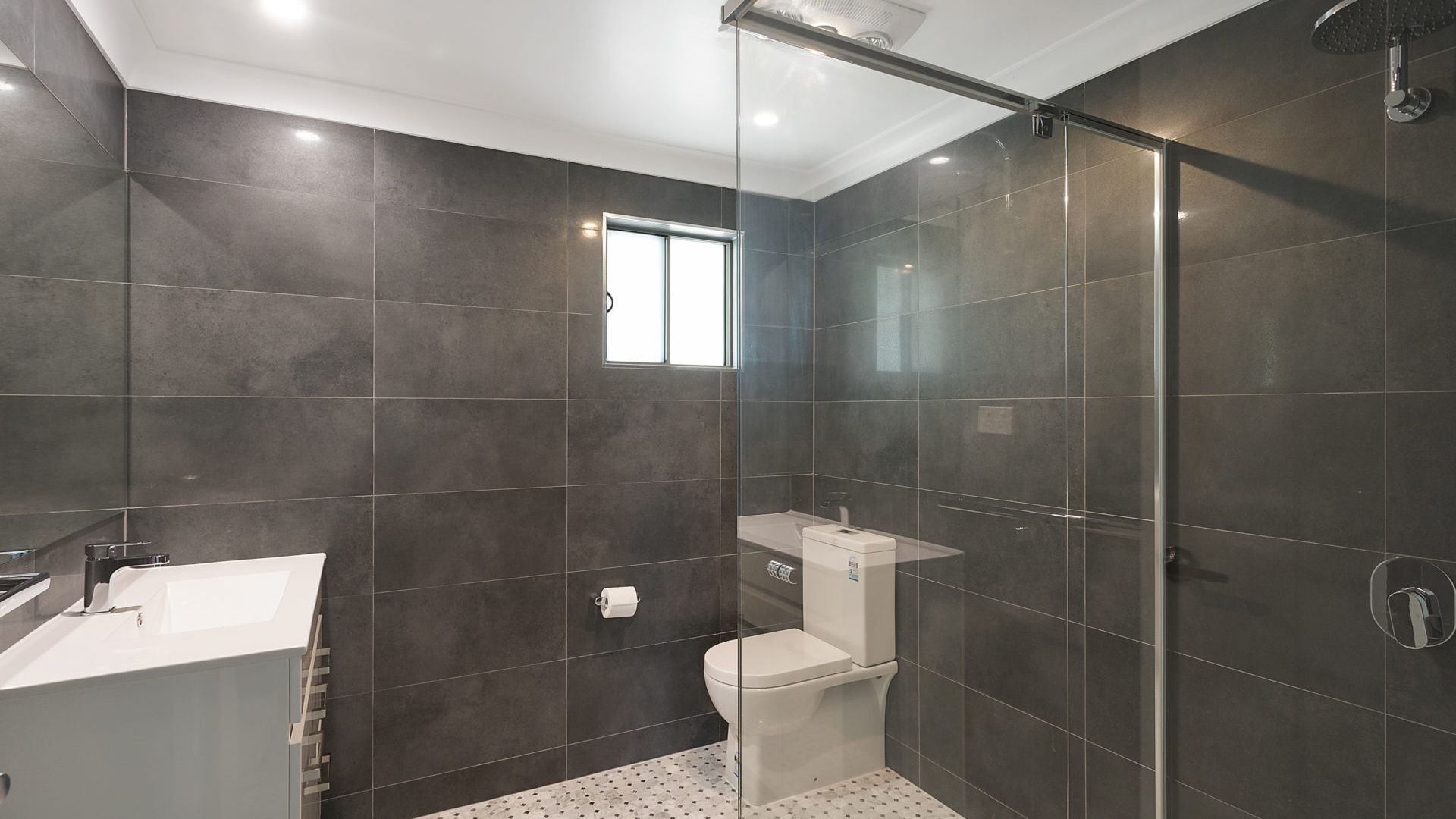 A modern bathroom with dark tiles, a glass shower enclosure, a white sink, and a toilet.