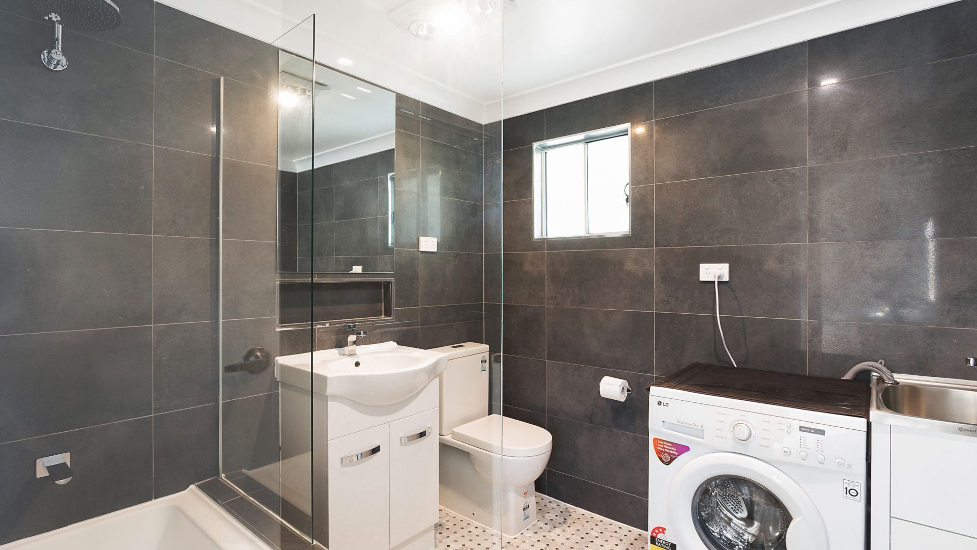 Une salle de bain moderne avec des carreaux foncés, une douche en verre, un lavabo blanc, des toilettes et un lave-linge à chargement frontal.