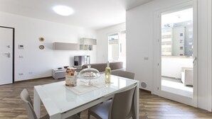 A modern, well-lit room with a dining table, chairs, a sofa, and a large window offering a view of the outside. - New and elegant central apartment with box (Alghero)