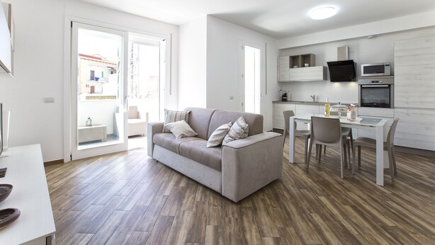A modern living room with a grey sofa, a dining table with chairs, a kitchen area with appliances, and a large window with a view of buildings outside. - New and elegant central apartment with box (Alghero)