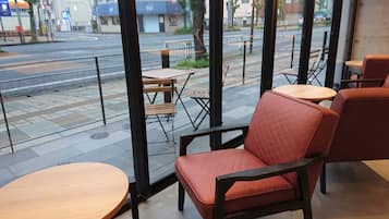 A cafe with red cushioned chairs and wooden tables, large windows with a view of a street and buildings.