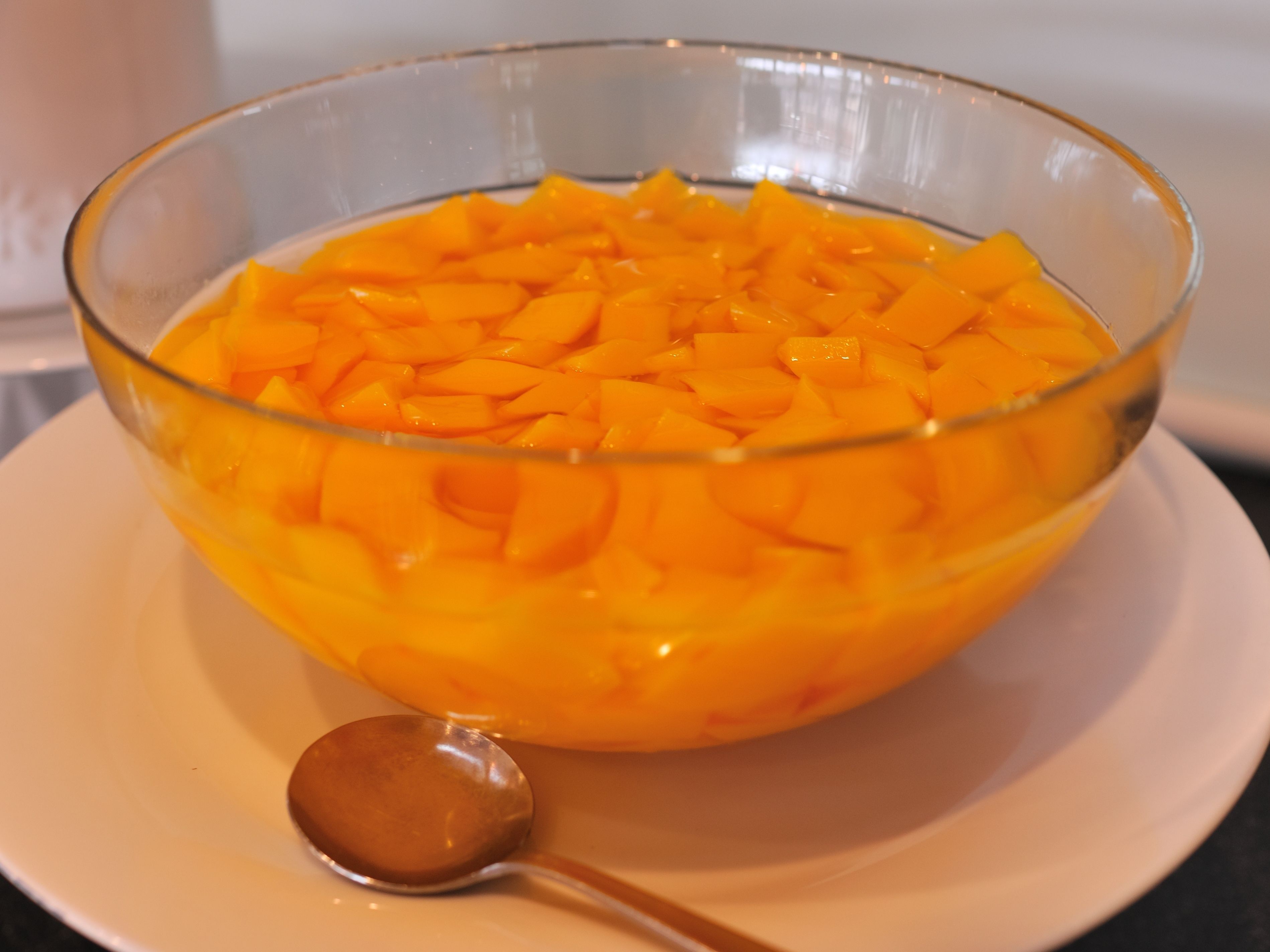 A clear glass bowl filled with diced mango on a white plate, accompanied by a spoon.