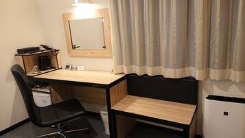 Une petite chambre d’hôtel avec un bureau en bois, une chaise de bureau noire, un petit réfrigérateur blanc, une machine à café et un miroir au mur.