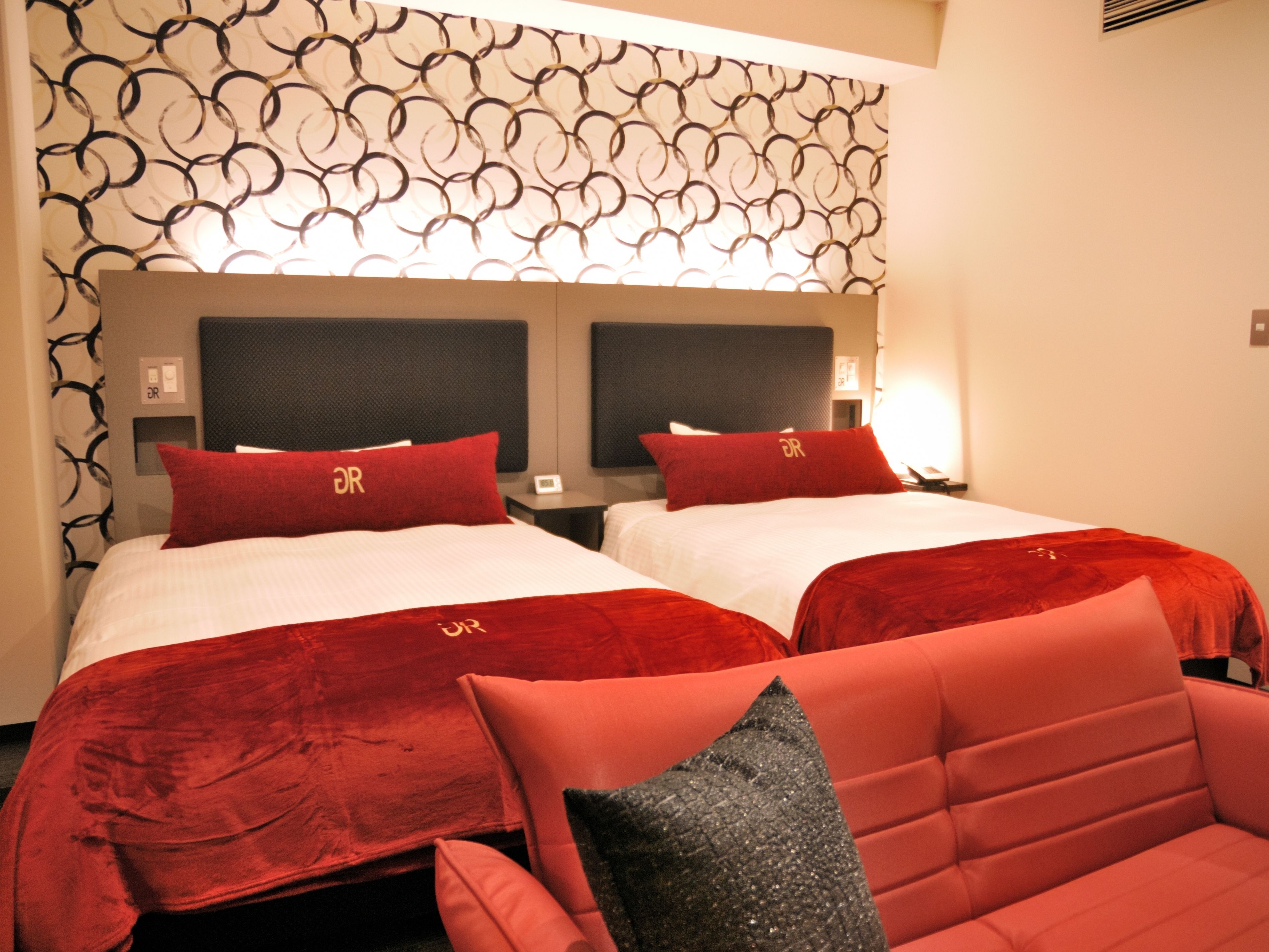 Habitación de hotel con dos camas, cada una con cabecero rojo y ropa de cama a juego. La habitación presenta papel tapiz estampado y un diseño moderno.