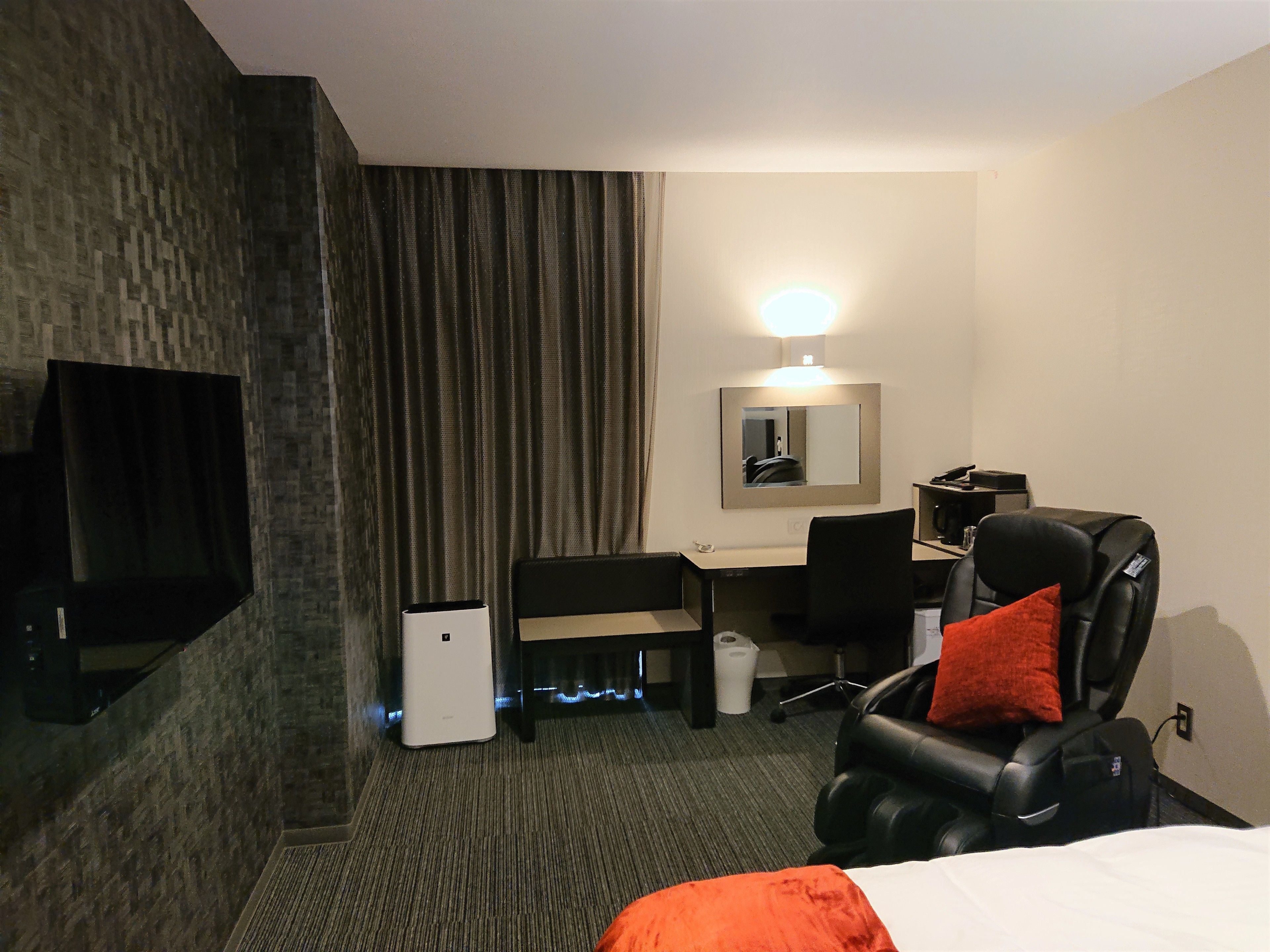 A hotel room with a bed, a desk, a chair, a massage chair, a small trash bin, and a flat-screen TV mounted on the wall.
