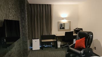 A hotel room with a bed, a desk, a chair, a massage chair, a small trash bin, and a flat-screen TV mounted on the wall.