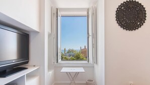 Een kleine kamer met een televisie op een standaard, een witte tafel en een raam met een open luik.