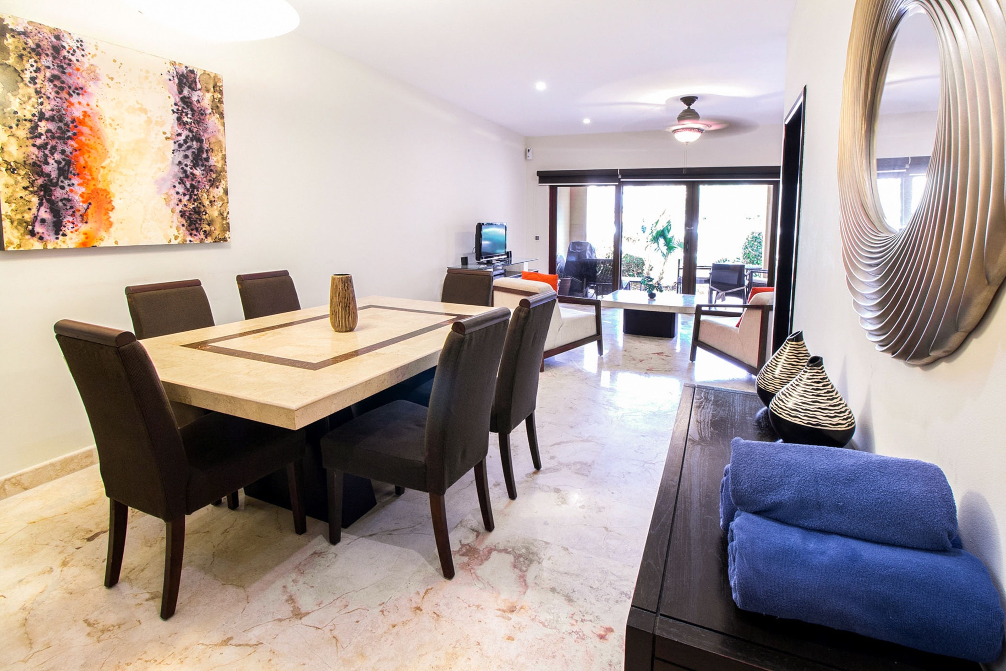 A modern dining area with a marble floor, a wooden dining table, and dark chairs. There is a television on a stand in the background, and a large abstract painting on the wall.