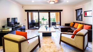 Una sala de estar con una mesa de centro de vidrio, dos sillones, un sofá con cojines coloridos, un televisor y una puerta corrediza de vidrio que da a un área exterior.