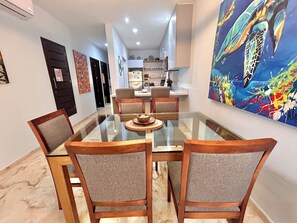 A dining area with a glass-top table surrounded by four chairs, a kitchen in the background, and a large abstract painting on the wall. - Sea Style Ocean View 2 Bedroom Condo, 2nd Floor at El Faro by DPM (Playa del Carmen)