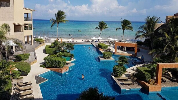A resort pool with a view of the beach and ocean, surrounded by palm trees and lounge chairs. - Sea Style Ocean View 2 Bedroom Condo, 2nd Floor at El Faro by DPM (Playa del Carmen)