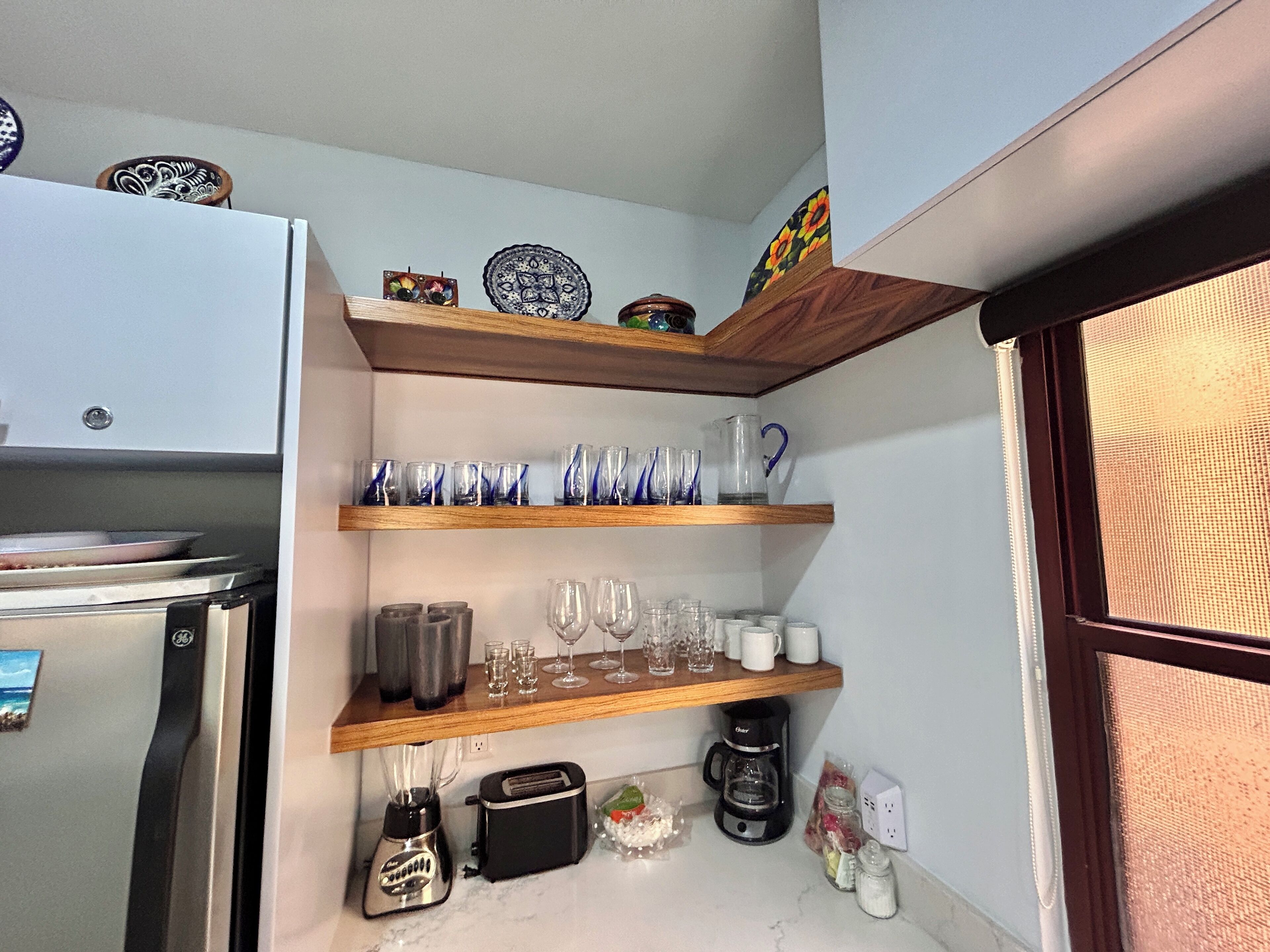 A kitchen with a refrigerator, a toaster, a blender, a coffee maker, and shelves with glassware and decorative items.