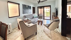 A living room with a sofa, armchairs, a coffee table, and a TV. There is a ceiling fan, a window, and a door leading outside. - Sea Style Ocean View 2 Bedroom Condo, 2nd Floor at El Faro by DPM (Playa del Carmen)