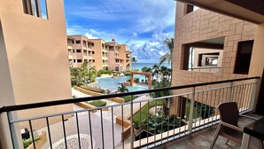 A balcony with a view of a swimming pool, palm trees, and a building with a red roof. - Sea Style Ocean View 2 Bedroom Condo, 2nd Floor at El Faro by DPM (Playa del Carmen)