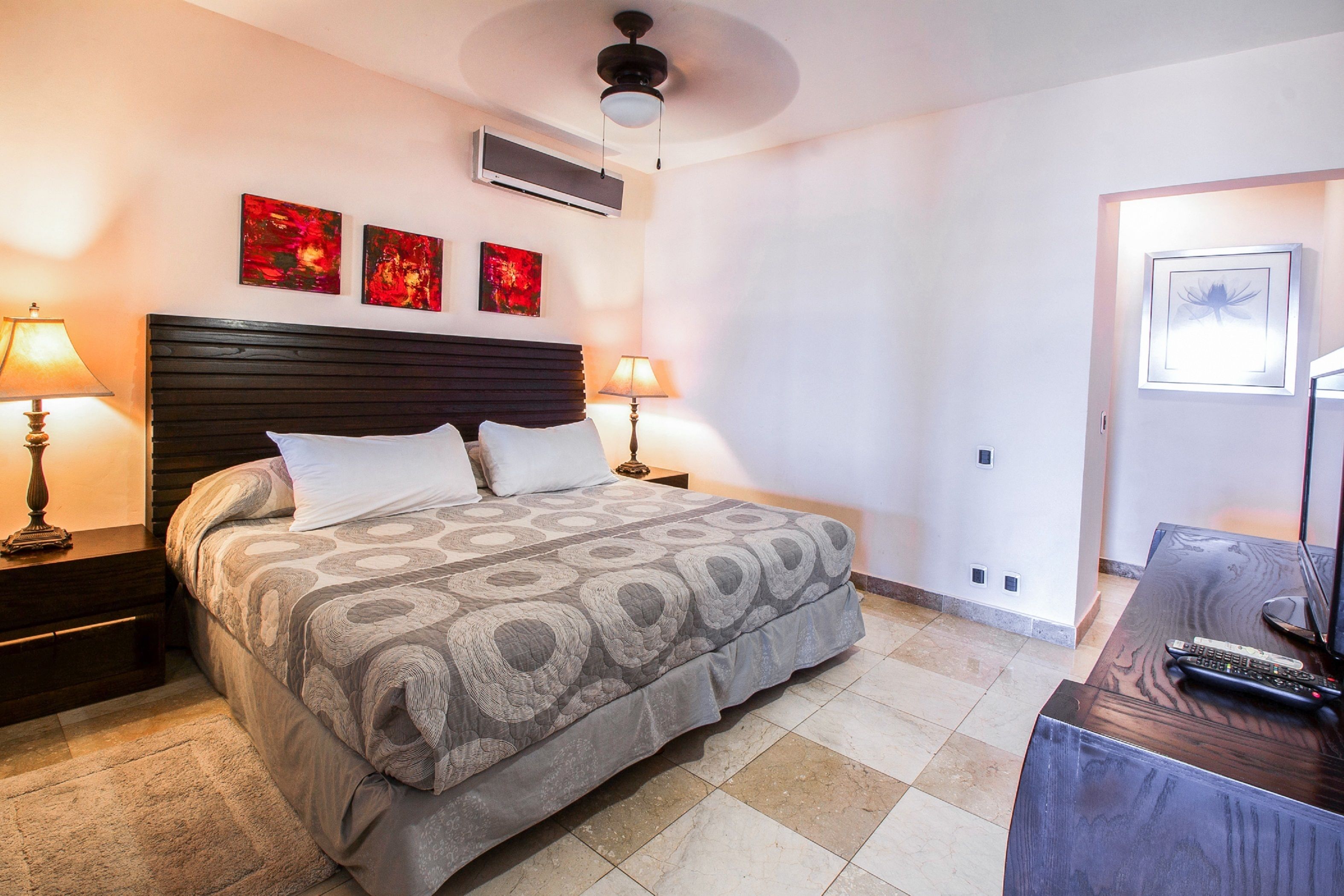 A hotel room with a bed, two bedside tables with lamps, a desk with a computer and keyboard, and artwork on the walls.
