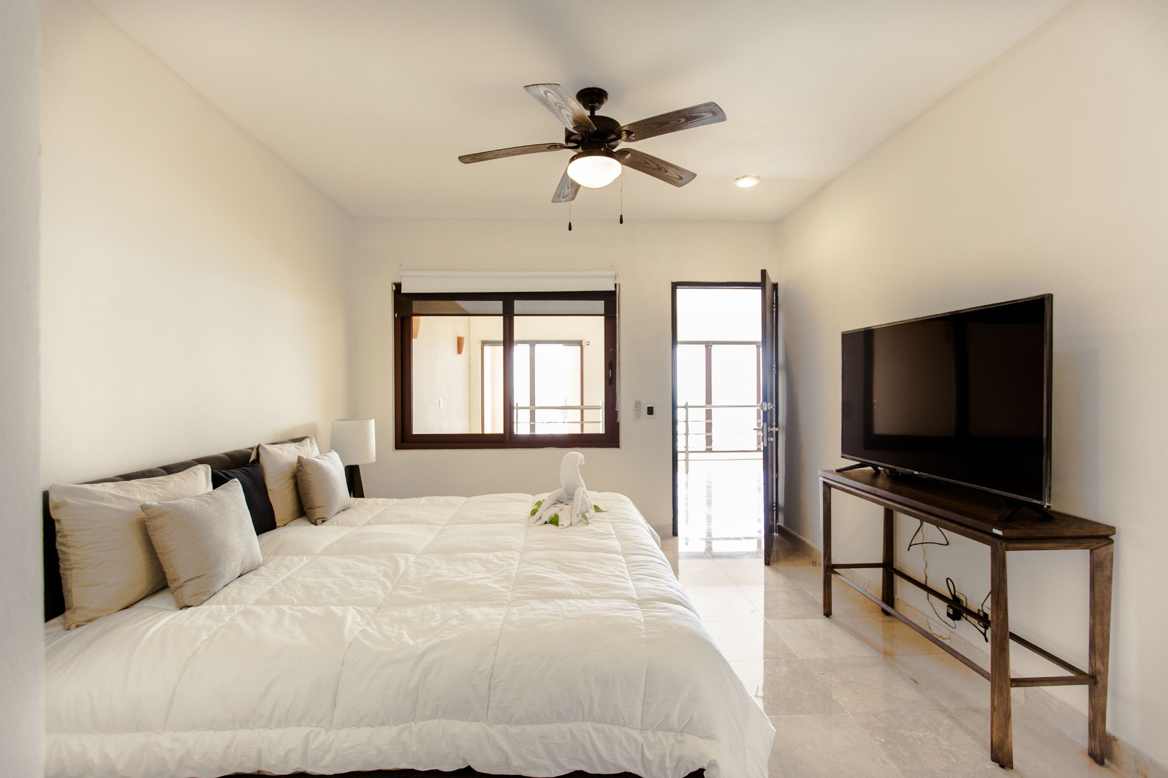 Une chambre à coucher avec un lit, un ventilateur de plafond, un téléviseur posé sur un meuble et une fenêtre.