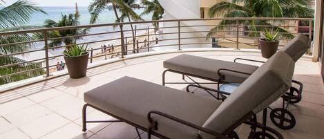 A balcony with two lounge chairs and potted plants, overlooking a beach with palm trees and a view of the ocean.