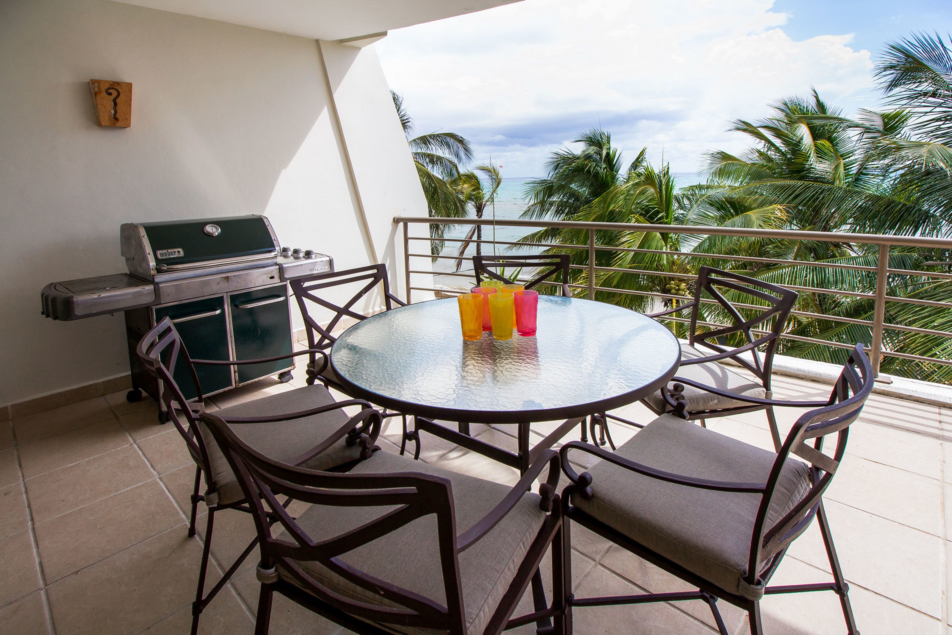 A round glass table with chairs on a balcony, a grill, and a view of the ocean.