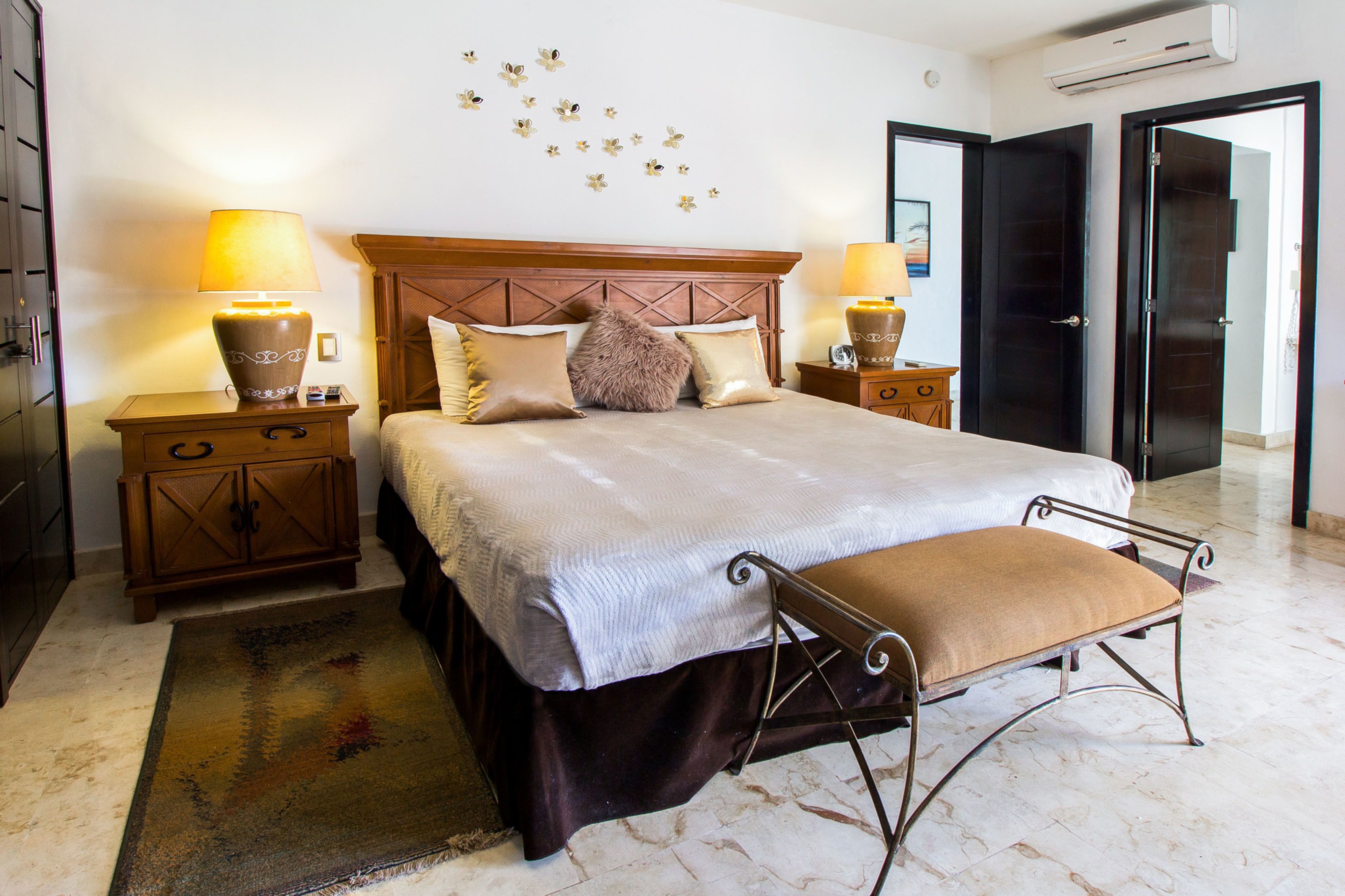 A hotel room with a large bed, two bedside tables with lamps, a wooden headboard, a bench at the foot of the bed, and a wall with decorative elements.