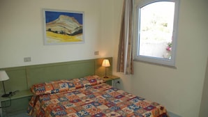 A bedroom with a bed, a nightstand, a lamp, a painting on the wall, and a window with curtains.