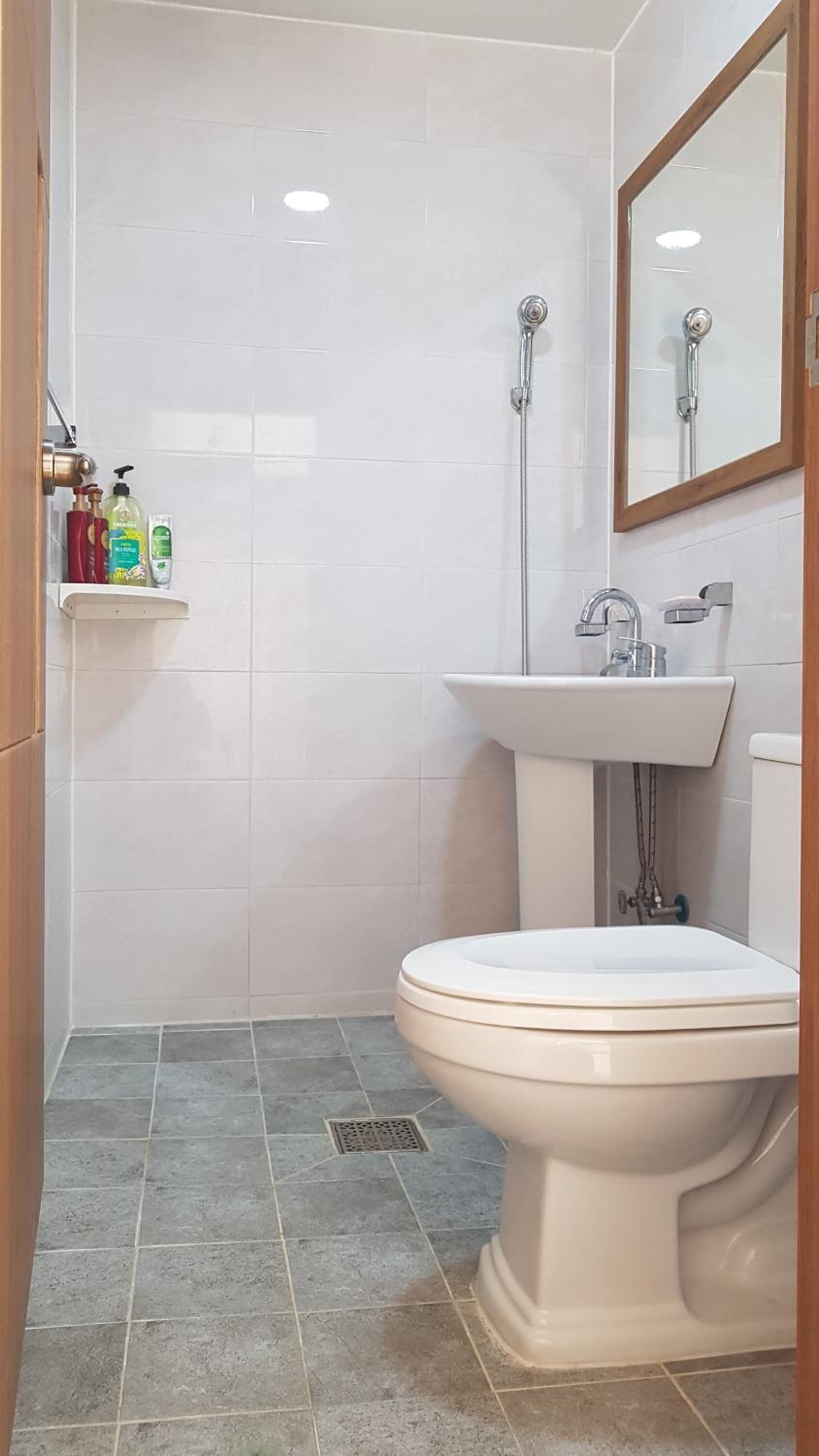 A bathroom with a toilet, sink, and shower. There is a shelf with toiletries.