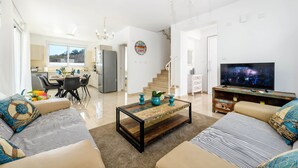 Een moderne woonkamer met een bank, salontafel en televisie. Er is een keukenhoek met een eettafel en stoelen. Achtergrond toont een trap.