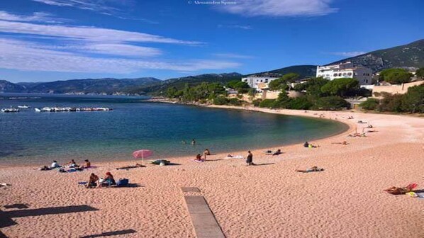 Una playa con gente tomando el sol y nadando, cielo despejado y edificios al fondo.