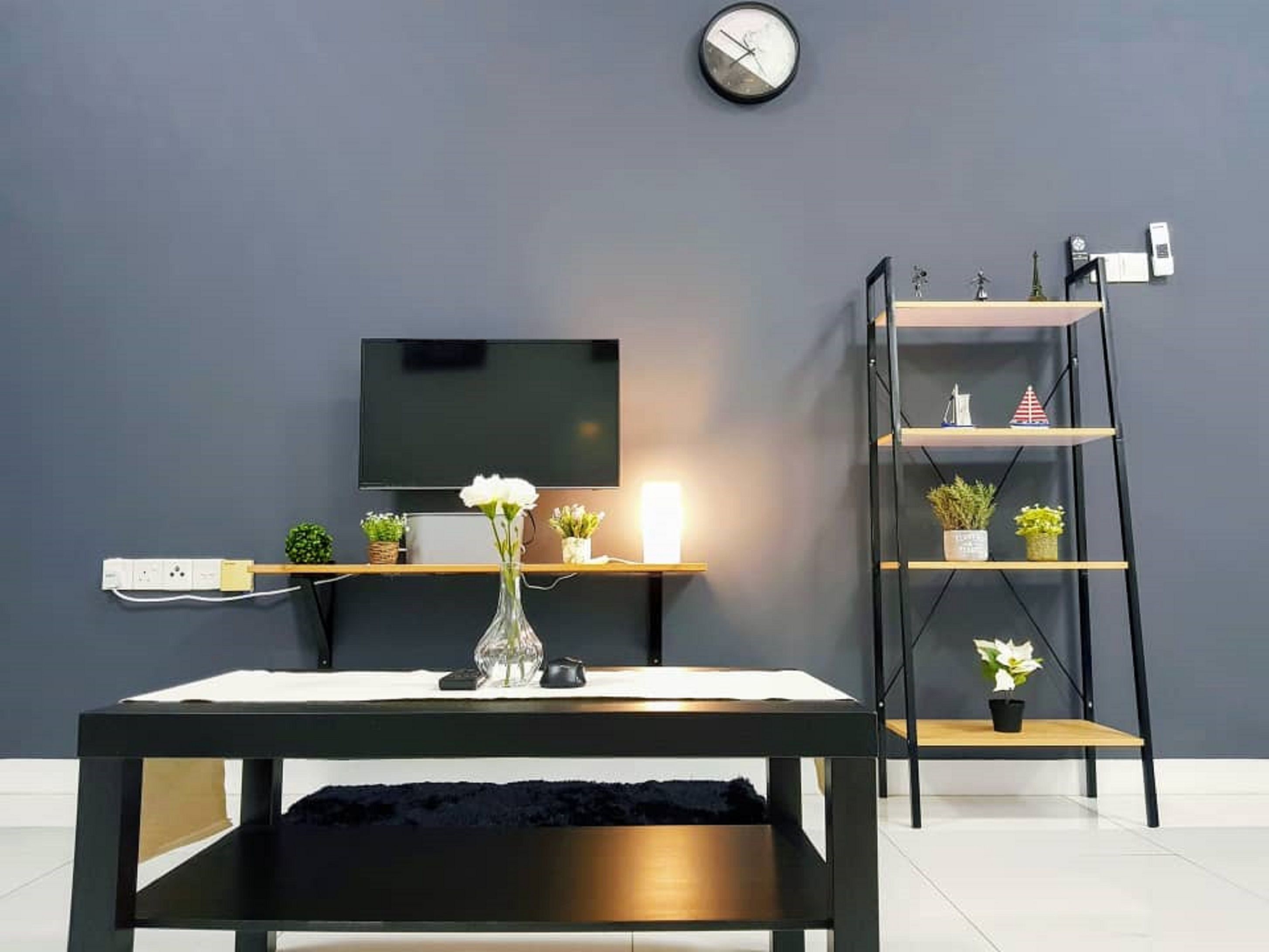 En moderne stue med et fladskærms-tv monteret på væggen, et træ sofabord med en vase med blomster, en sort bogreol med pynteting, og en sort sofa.