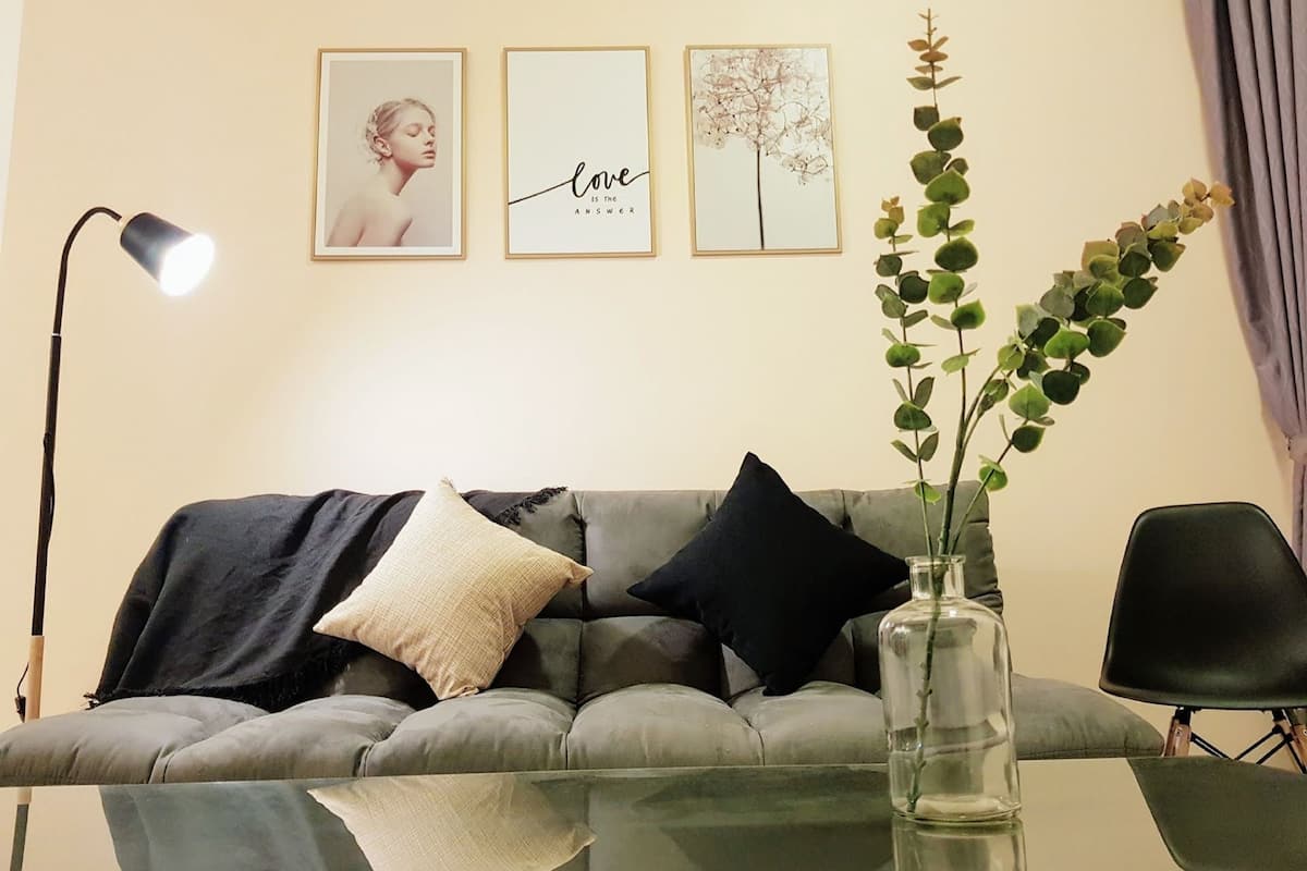 Ein modernes Wohnzimmer mit einem grauen Sofa, einem Glastisch und einer Vase mit Grünpflanzen. An der Wand hängen drei gerahmte Bilder, eines mit der Aufschrift 'love is the answer'.