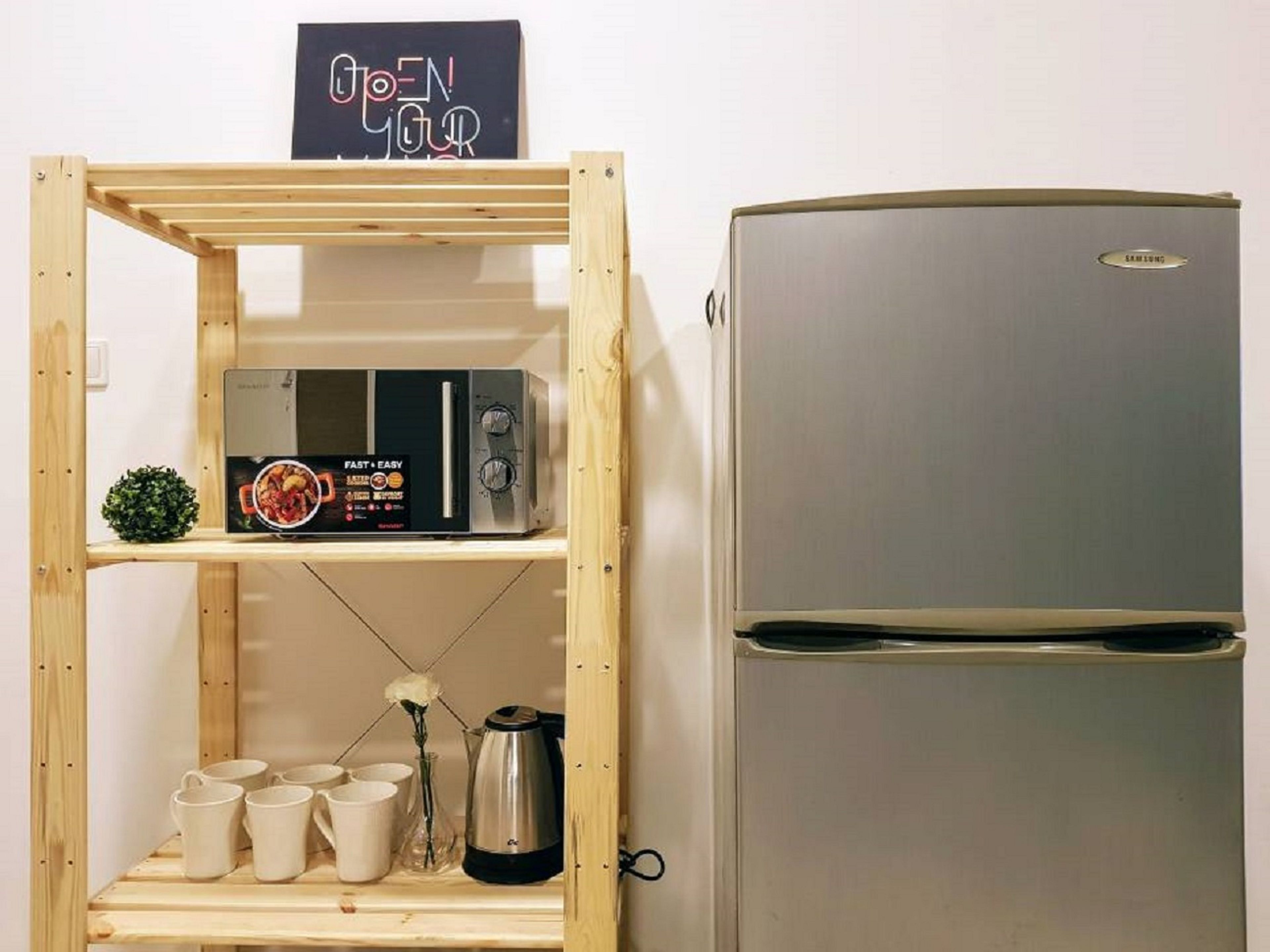 A kitchen with a stainless steel refrigerator, a microwave on a wooden shelf, and a sign that says "OPEN YOUR REFRIGERATOR".