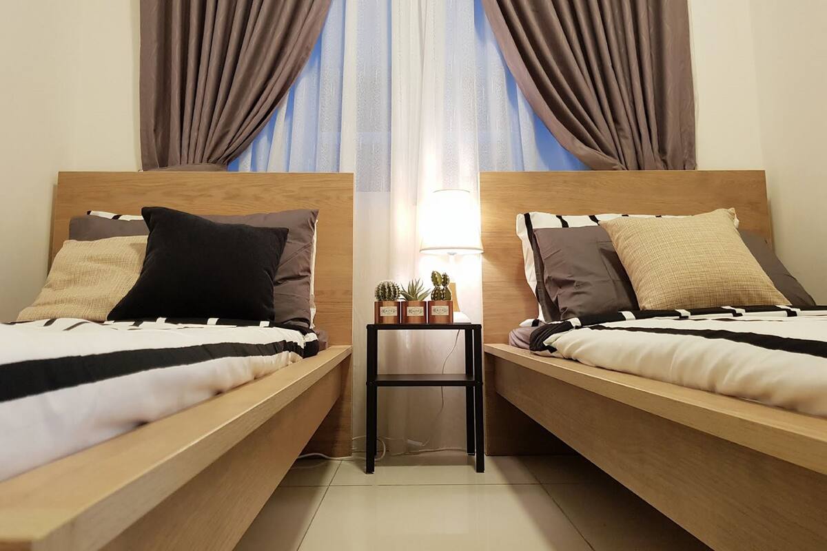 Ein Hotelzimmer mit zwei Holzbettgestellen, einem Nachttisch mit Lampe und Blick durch transparente Vorhänge.