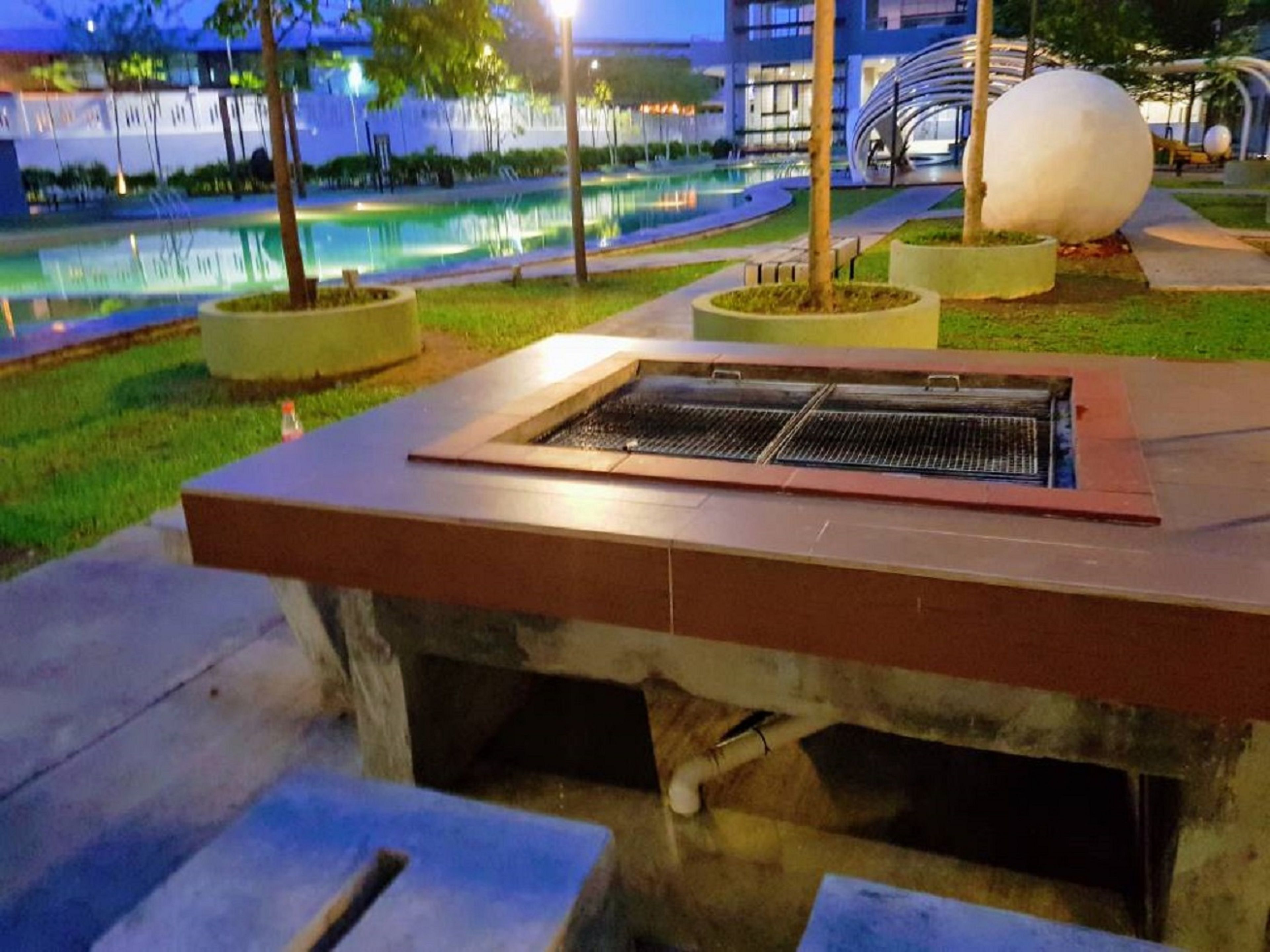 Ein Grillbereich im Freien mit einer Steinsitzbank, umgeben von einem angelegten Garten mit Bäumen und einem Wasserspiel.