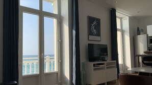 A modern living room with a large window overlooking the sea, a flat-screen TV on a white cabinet, and a dining area with a table and chairs.
